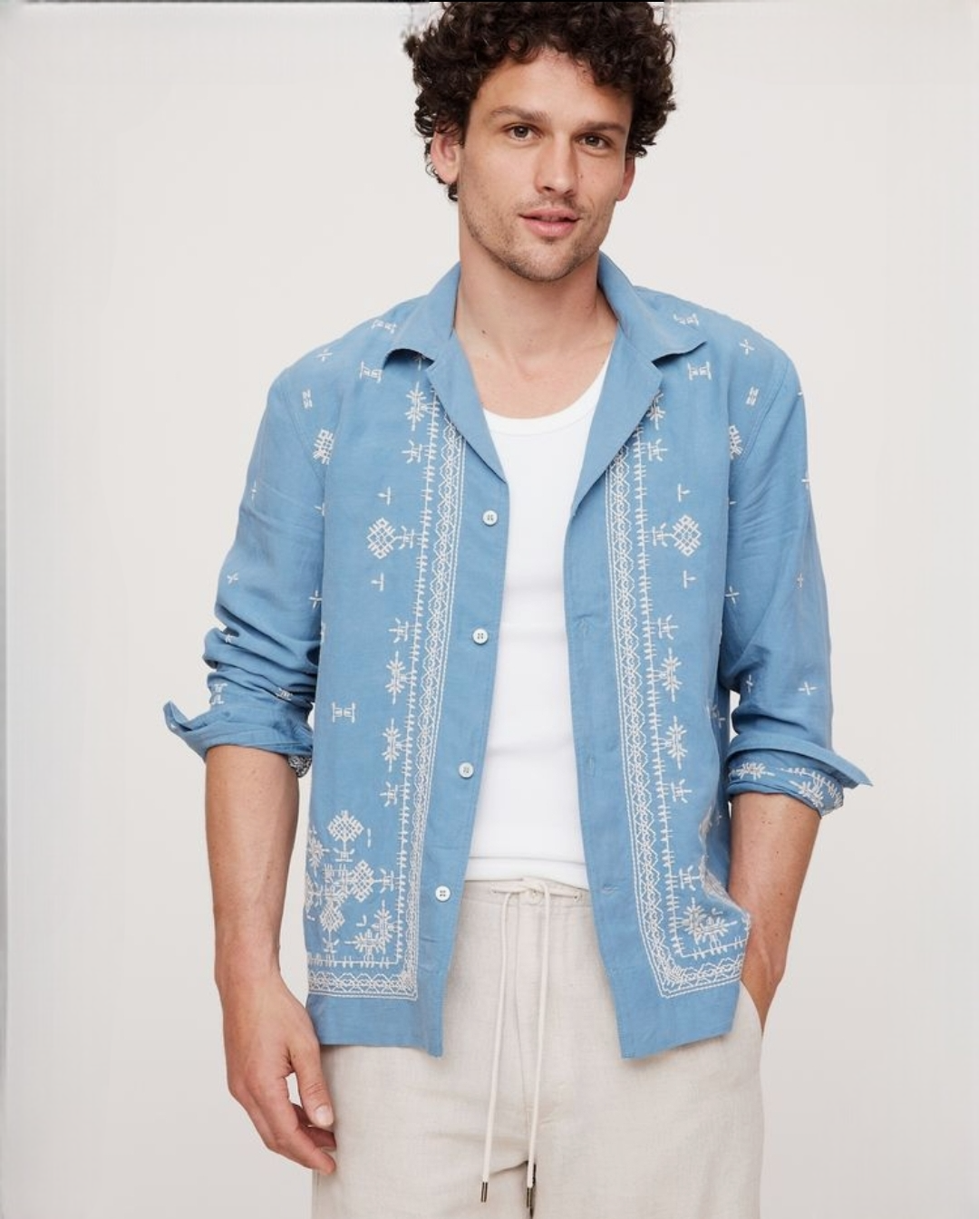 Man wearing a blue embroidered shirt over a white t-shirt on a plain background